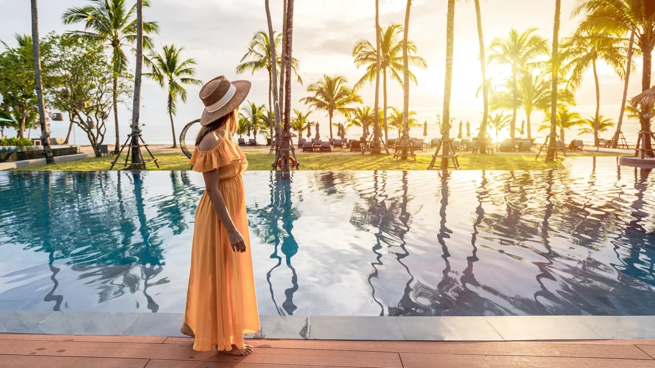 A luxury vacation scene of a woman in an orange dress standing next to a pool as she looks towards the ocean and palm trees.