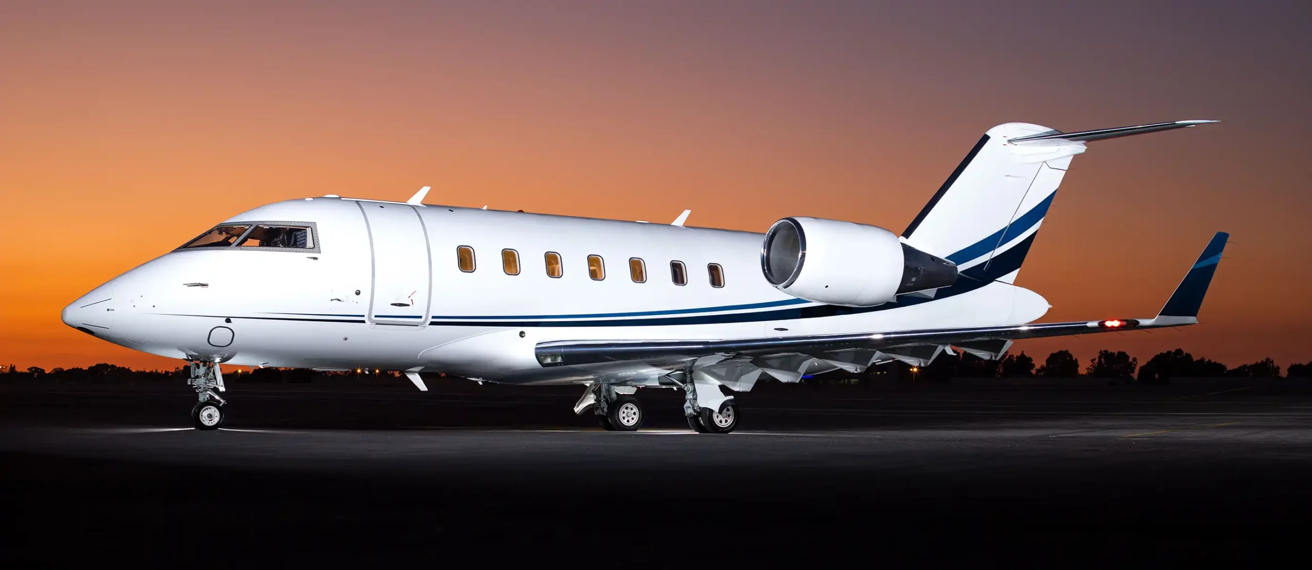 A Latitude 33 Aviation private jet with a white exterior and blue details sits on the runway as the sun sets or rises.