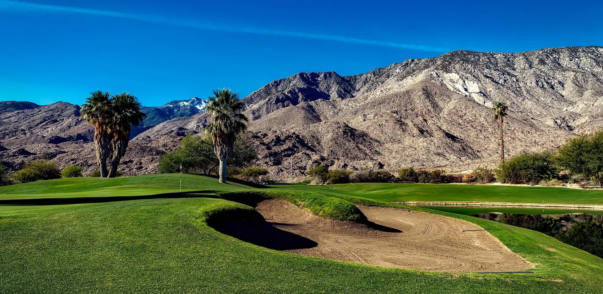 A photo of a green at the Indian Canyons golf resort in Palm Springs, CA. Desert mountains, palm trees, a lake, and a bunker fill the scene.
