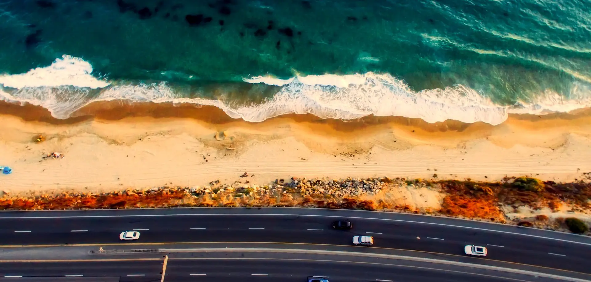 A top-down view of the Laguna Beach shoreline. The deep, greenish-blue sea crashes onto the beach. A highway crosses the bottom third of the photo.