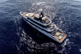 Aerial view of motor yacht Cada Dia cruising across deep blue water, showcasing her expansive decks, sleek profile, and private beach club with spa pool.